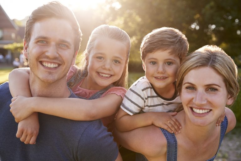 Portrait Of Smiling Family Outdoors In Summer Park Against Flaring Sun